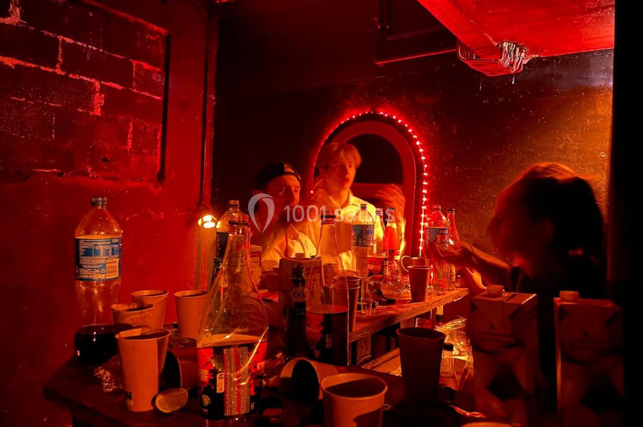 Des personnes discutent autour d'une table encombrée de bouteilles et gobelets dans une pièce éclairée par une lumière rouge.
