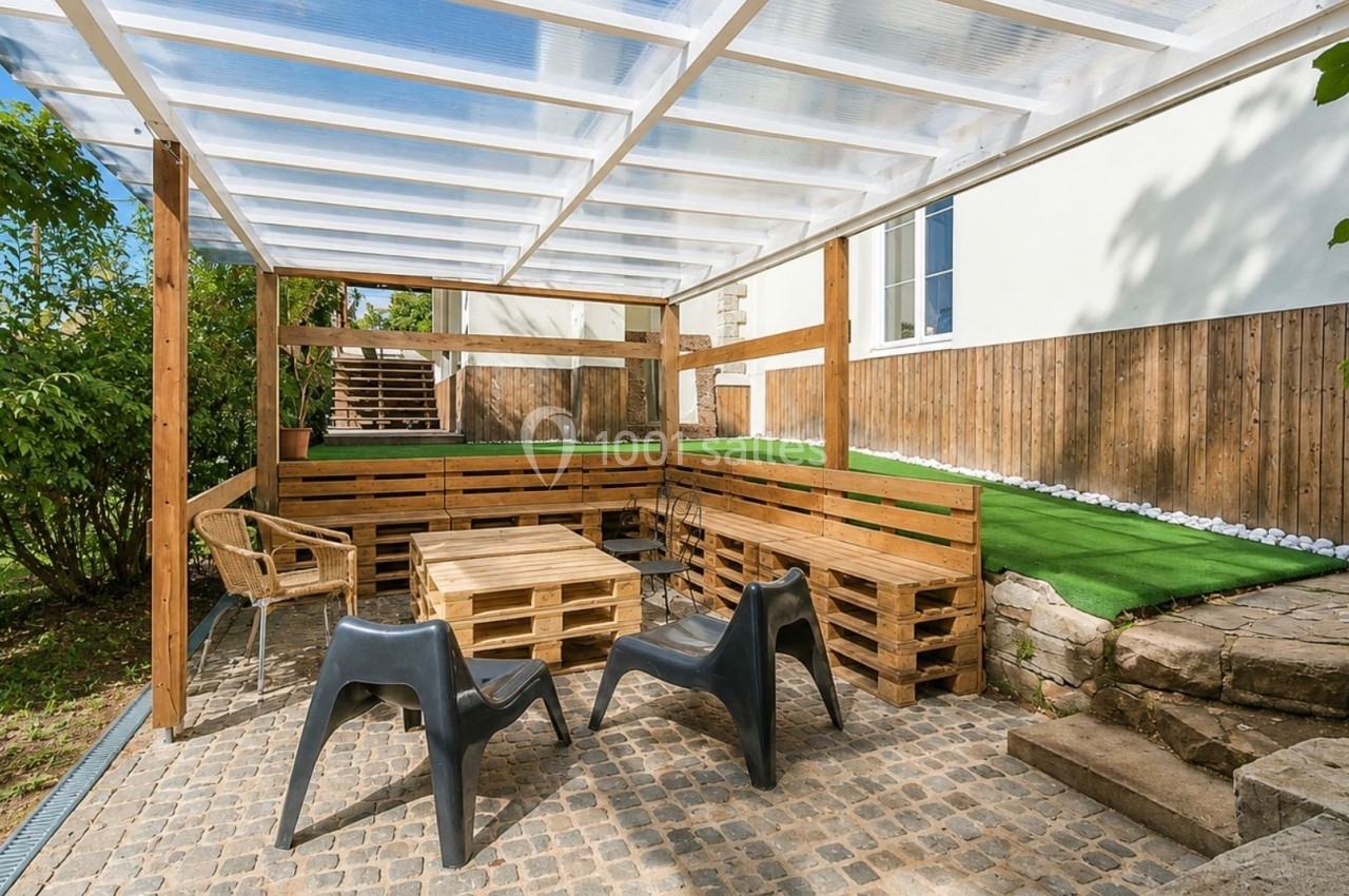 Terrasse extérieure avec mobilier en palettes, chaises en plastique, sol pavé et pergola en toit transparent.