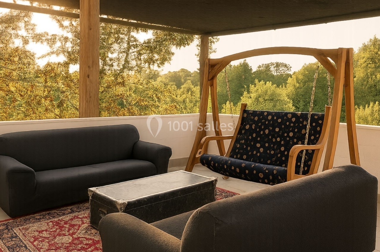 Terrasse avec canapé, fauteuil suspendu en bois, table basse et vue sur des arbres au coucher du soleil.