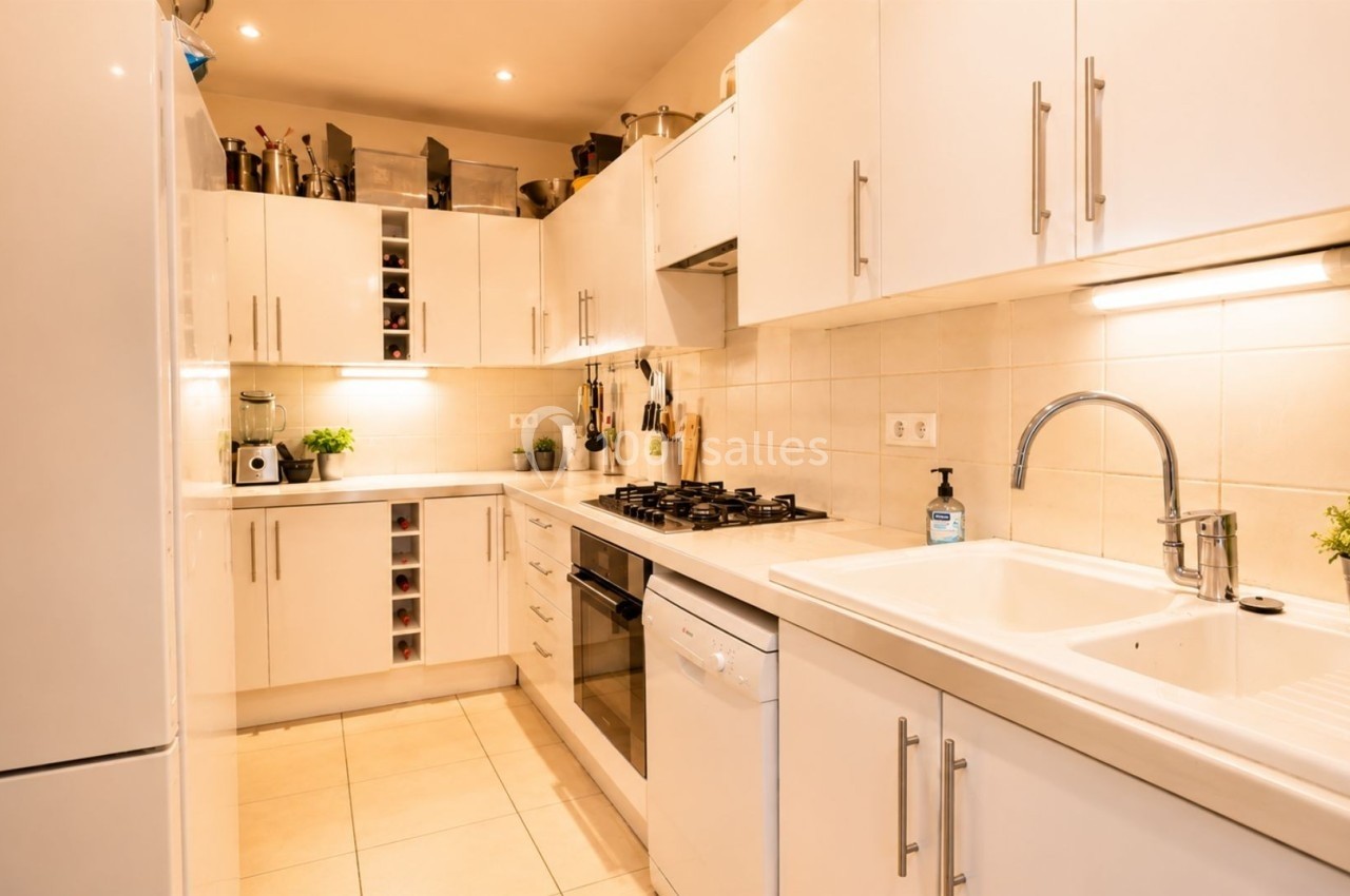 Cuisine moderne avec meubles blancs, évier en inox, plaques de cuisson, réfrigérateur et éclairage chaleureux.