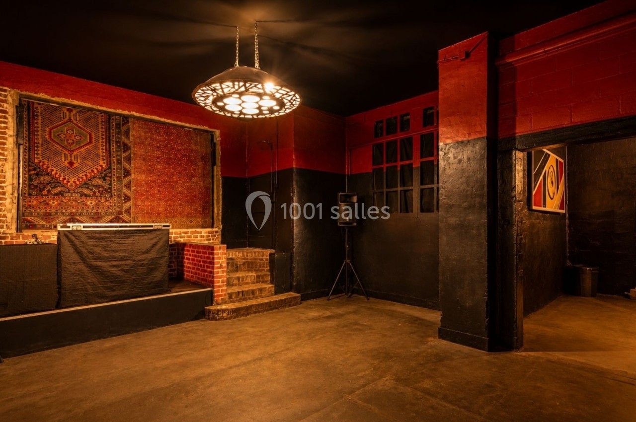 Salle vide avec murs rouges et noirs, tapis mural, éclairage suspendu, estrade avec table de mixage et enceintes.