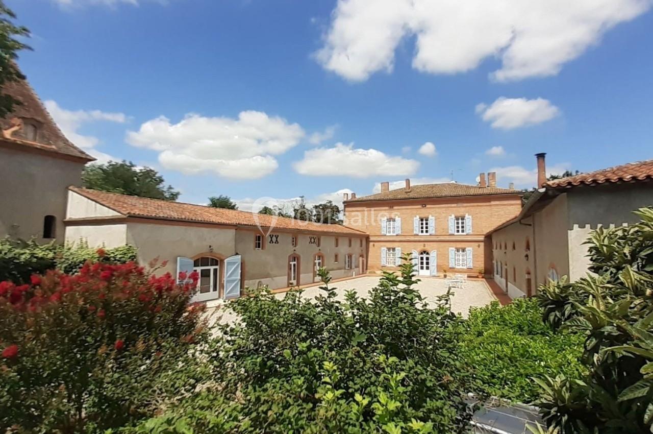 Cour intérieure d'un domaine avec bâtiments en pierre, végétation au premier plan et ciel bleu parsemé de nuages.