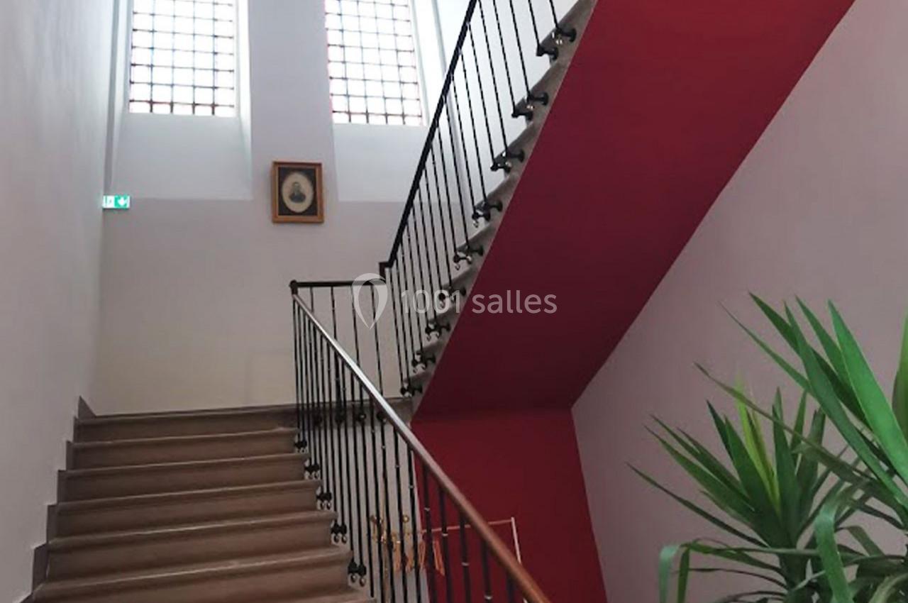 Escalier intérieur en bois avec rampe métallique, murs blancs et rouges, grandes fenêtres en arrière-plan.