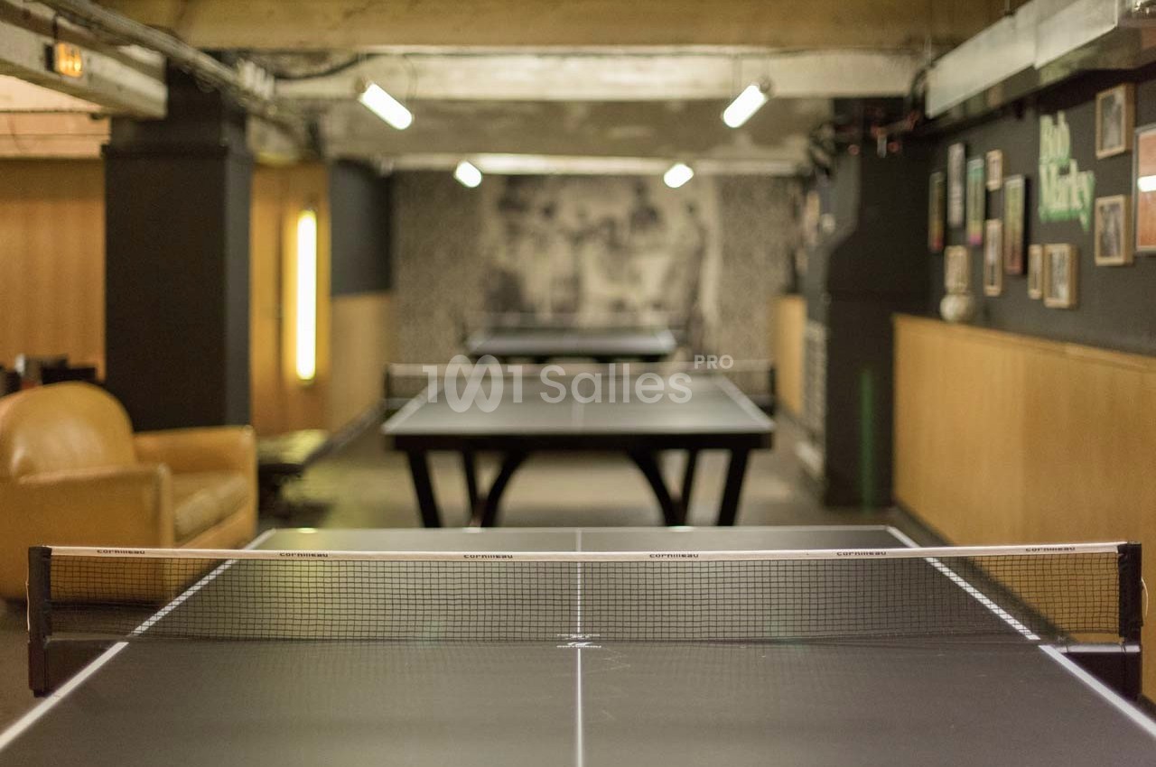 Salle intérieure avec plusieurs tables de ping-pong alignées sous un éclairage tamisé.
