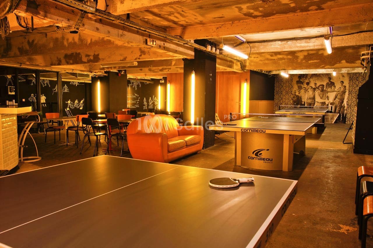 Salle de loisirs avec tables de ping-pong, canapé orange, chaises et éclairage chaleureux dans un espace industriel.