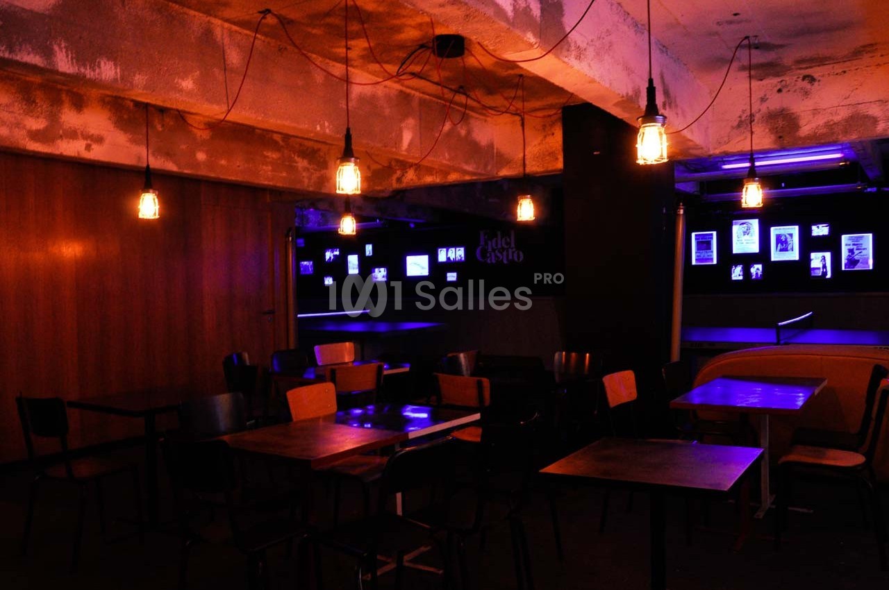 Salle sombre avec des tables et chaises vides, éclairée par des ampoules suspendues et des lumières violettes.