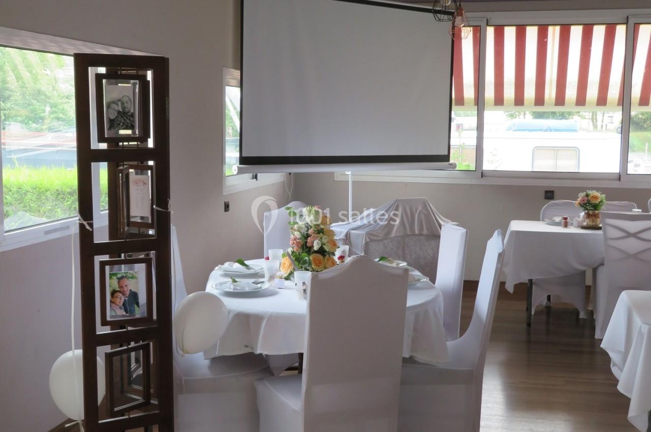 Salle de réception décorée avec des tables rondes, nappes blanches et un bouquet de fleurs au centre.