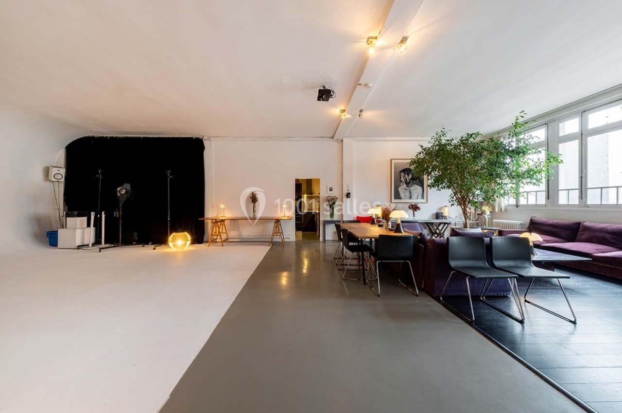 Salle lumineuse avec espace studio photo, coin salon avec canapé, table à manger et grande plante décorative.