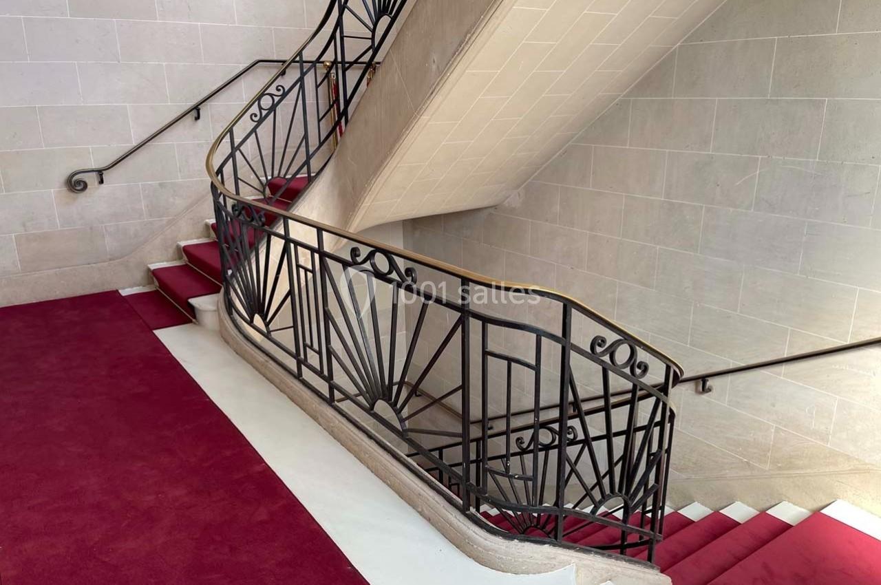 Escalier intérieur en pierre avec rampe en fer forgé décoratif et tapis rouge sur les marches.