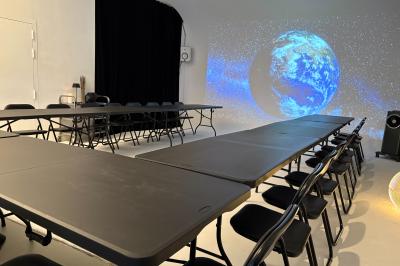 Miniature Location salle Paris 20 (Paris) - Studio 7 #18 Salle avec des tables et chaises noires disposées en U, projection d’un paon coloré sur un mur blanc, rideaux noirs à droite.