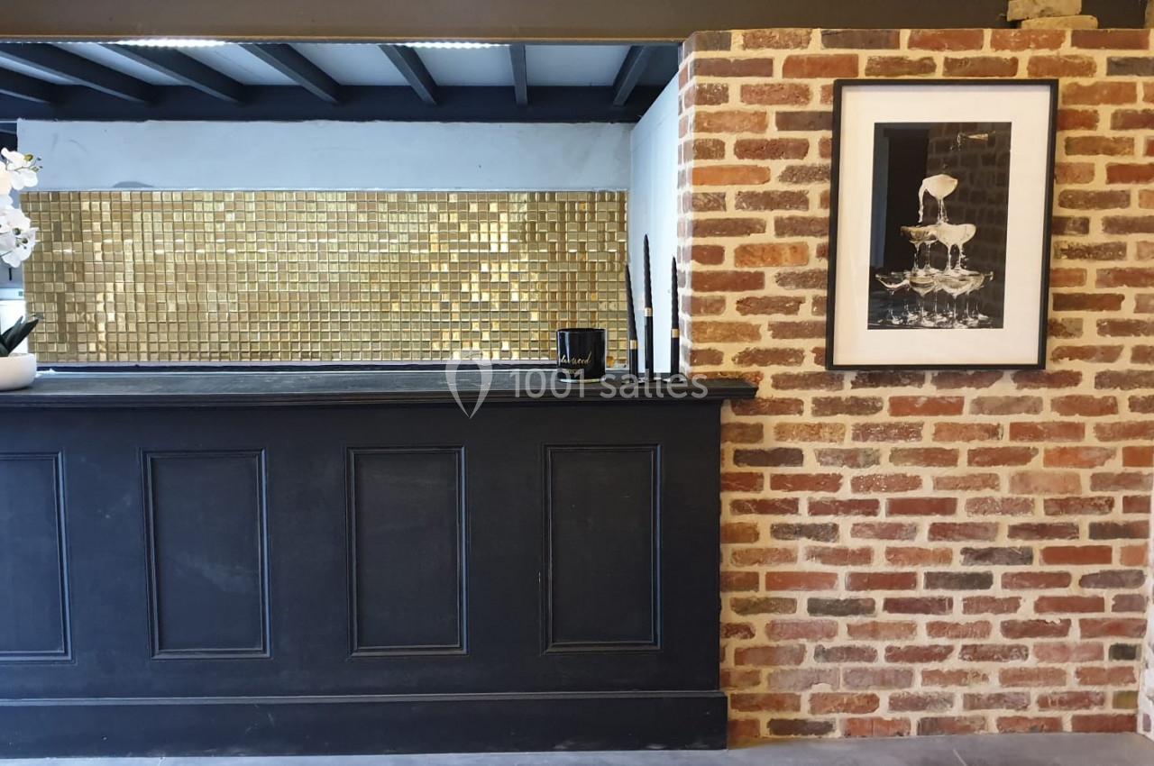 Location salle Haverskerque (Nord) - Ferme d'Hercule #10 Comptoir noir devant un mur en briques, décoré d'un cadre photo, d'une orchidée blanche et d'un fond doré.