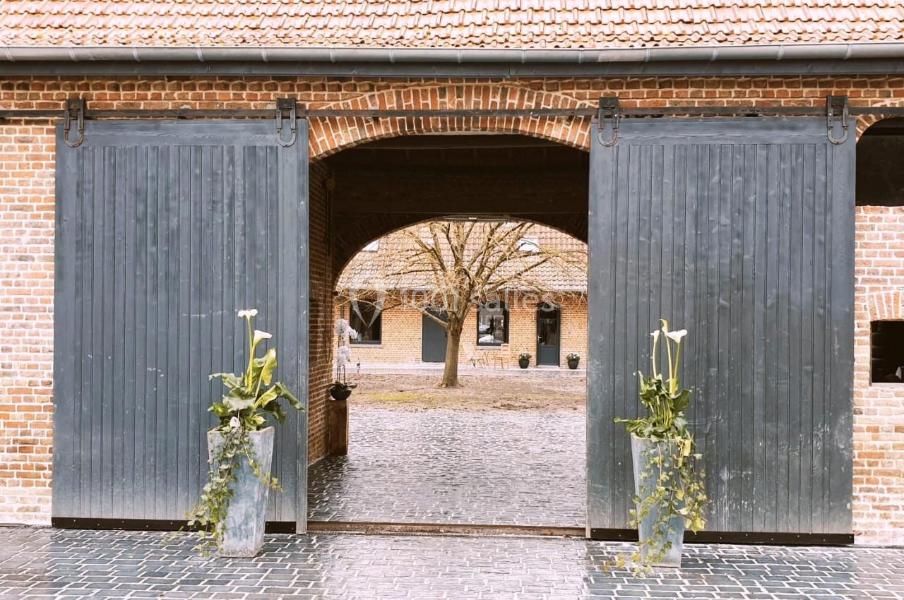 Location salle Haverskerque (Nord) - Ferme d'Hercule #13 Portail en bois gris ouvert sur une cour pavée, entourée de bâtiments en briques avec des plantes décoratives.