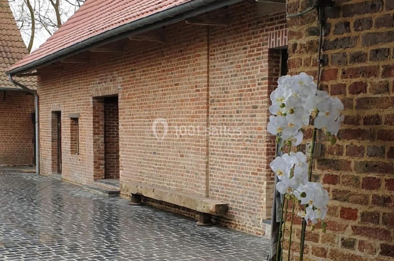 Location salle Haverskerque (Nord) - Ferme d'Hercule #12 Cour pavée bordée de bâtiments en briques rouges, avec une orchidée blanche en premier plan.