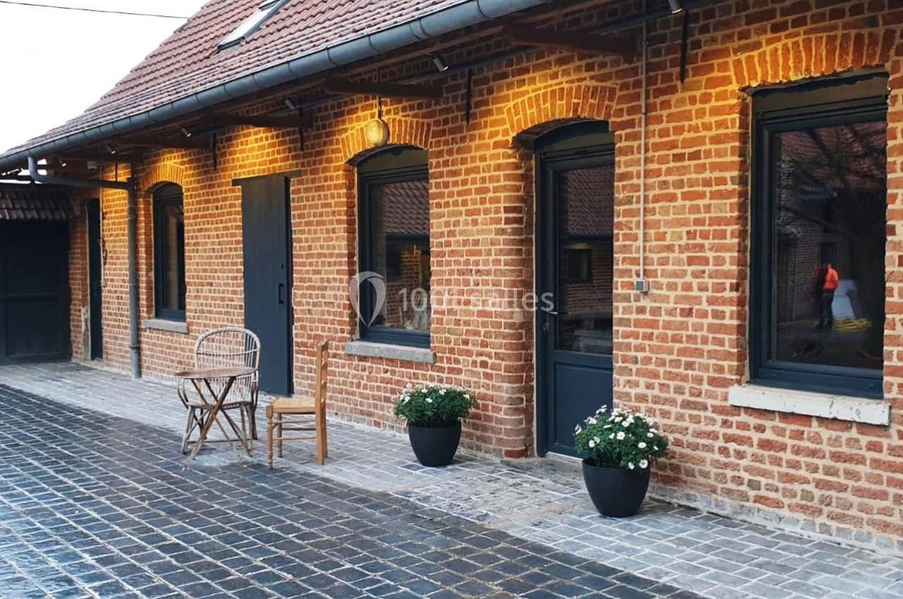 Location salle Haverskerque (Nord) - Ferme d'Hercule #8 Façade en briques rouges avec fenêtres noires, deux chaises en bois, pots de fleurs et pavés au sol.