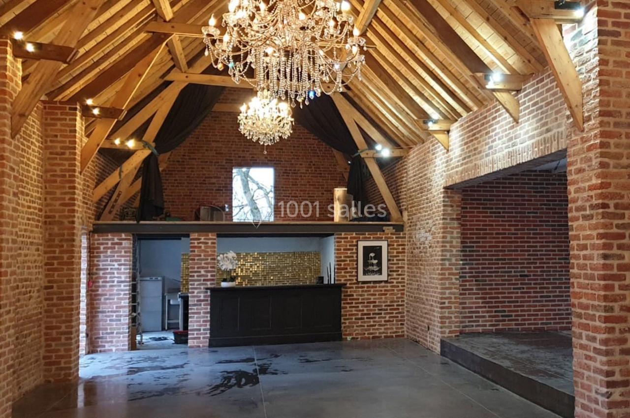 Location salle Haverskerque (Nord) - Ferme d'Hercule #7 Intérieur d'une salle avec murs en briques, plafond en bois, lustres suspendus et une mezzanine au fond.