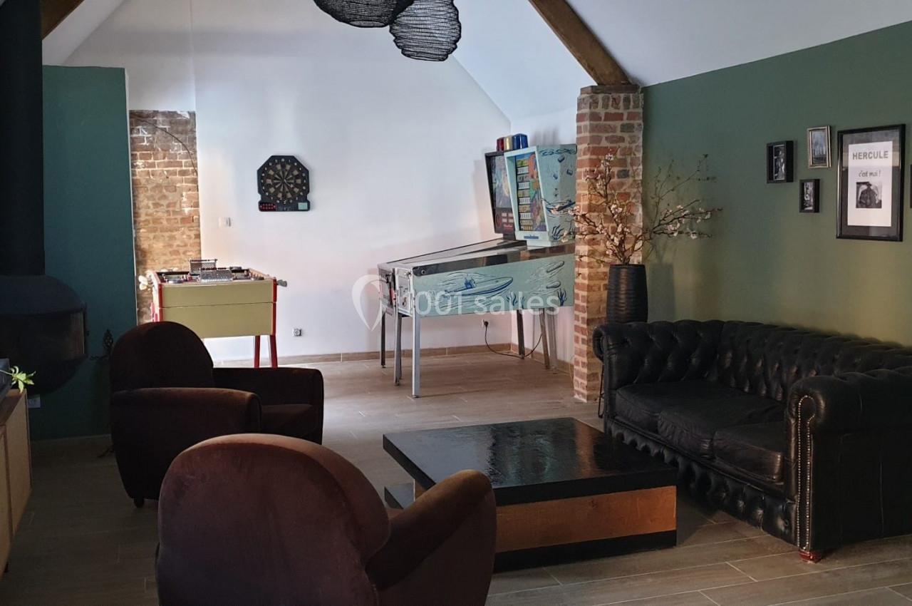 Location salle Haverskerque (Nord) - Ferme d'Hercule #22 Salon aménagé avec canapé en cuir, fauteuils, poêle, baby-foot, flipper et murs verts décorés de cadres.