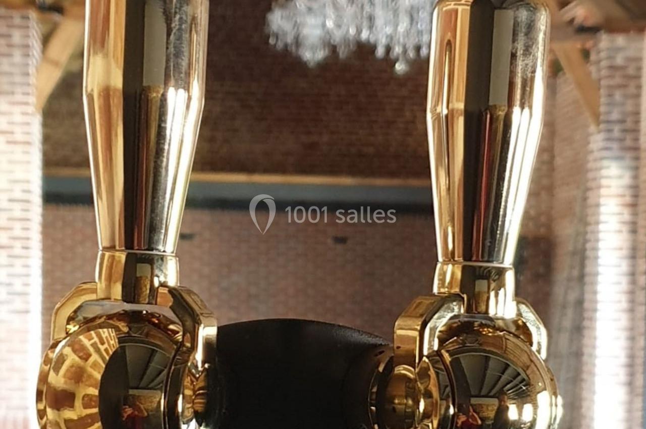 Location salle Haverskerque (Nord) - Ferme d'Hercule #28 Deux robinets dorés brillants sur une tireuse à bière, avec un arrière-plan en briques et un lustre suspendu.
