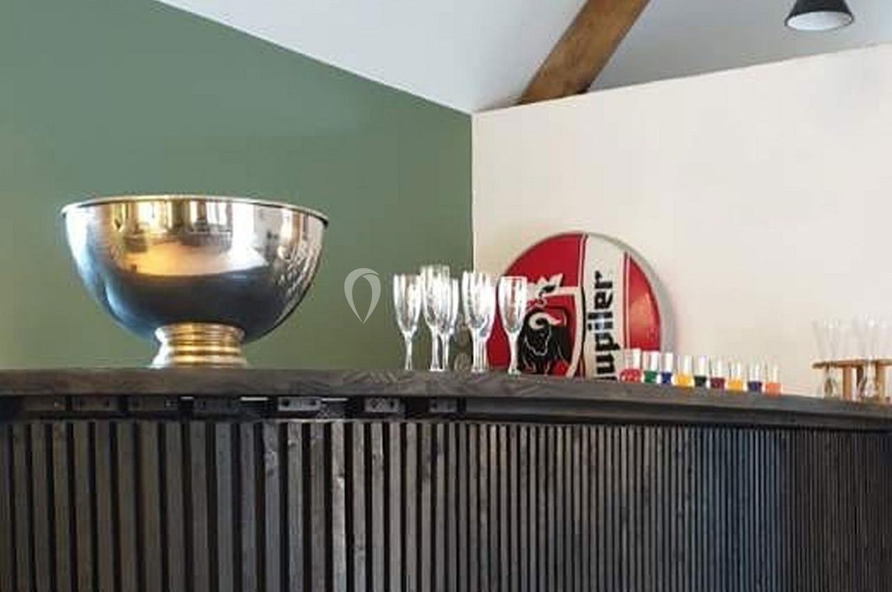 Location salle Haverskerque (Nord) - Ferme d'Hercule #23 Un comptoir en bois sombre avec une grande coupe argentée, des verres et un panneau décoratif en arrière-plan.