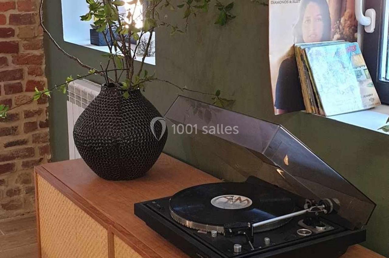 Location salle Haverskerque (Nord) - Ferme d'Hercule #20 Platine vinyle sur un meuble en bois, avec un vase noir et des branches, près d'une fenêtre et d'une affiche.
