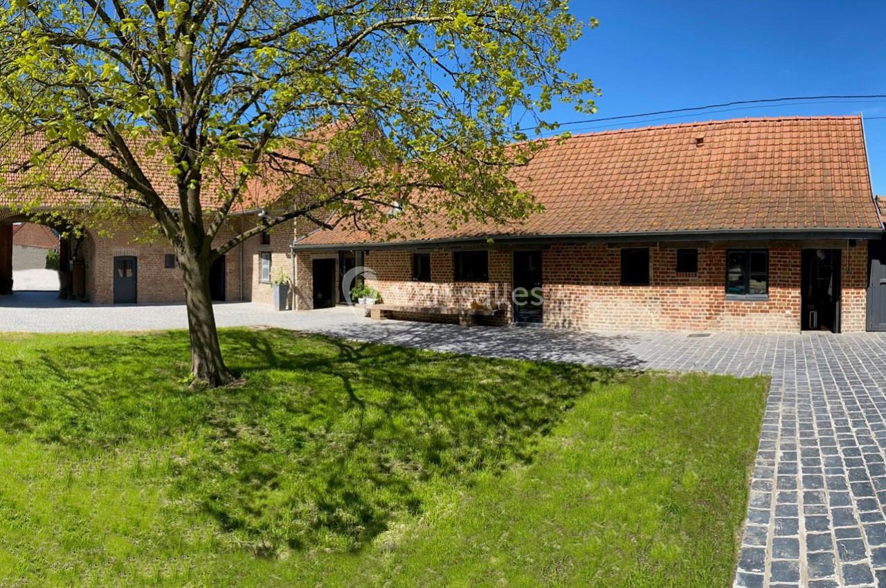 Location salle Haverskerque (Nord) - Ferme d'Hercule #30 Bâtiment en briques rouges avec toit en tuiles, cour pavée, pelouse et arbre sous un ciel bleu.