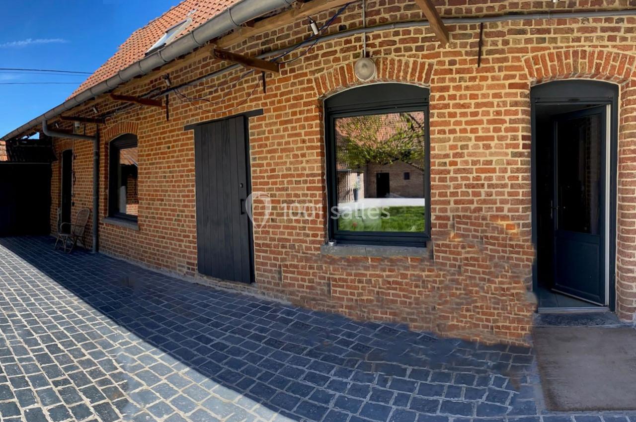 Location salle Haverskerque (Nord) - Ferme d'Hercule #32 Façade en briques rouges avec portes et fenêtres noires, donnant sur une cour pavée ensoleillée.