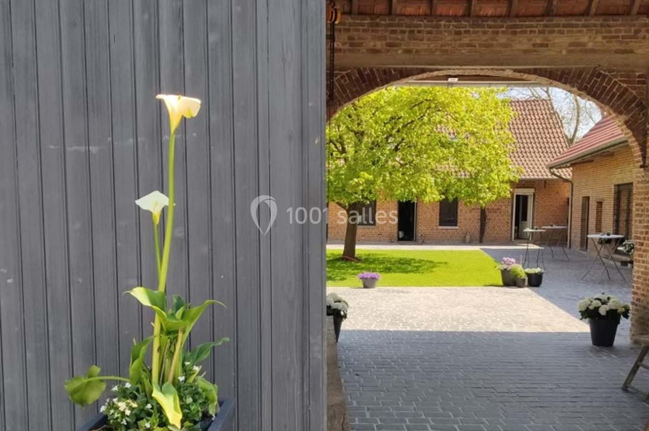 Location salle Haverskerque (Nord) - Ferme d'Hercule #29 Plante avec des arums blancs en pot devant une porte grise, ouvrant sur une cour pavée et un arbre verdoyant.