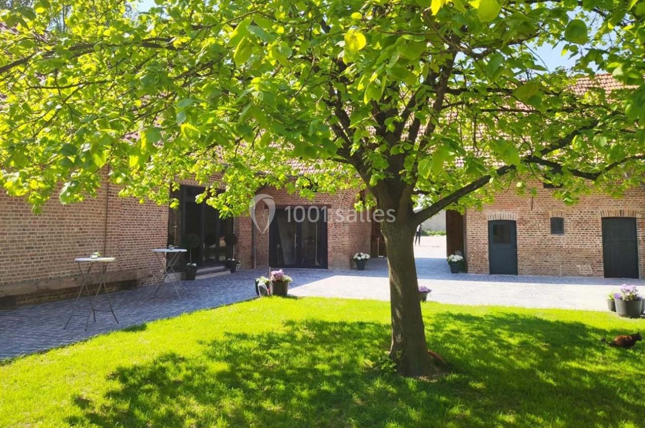 Location salle Haverskerque (Nord) - Ferme d'Hercule #31 Cour pavée avec un arbre au centre, entourée de bâtiments en briques et de pots de fleurs sous un ciel ensoleillé.