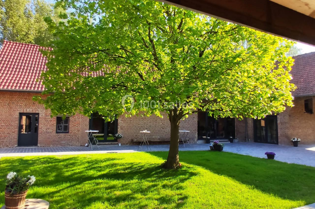 Location salle Haverskerque (Nord) - Ferme d'Hercule #33 Cour ensoleillée avec un arbre central, entourée de bâtiments en briques et de mobilier de jardin.