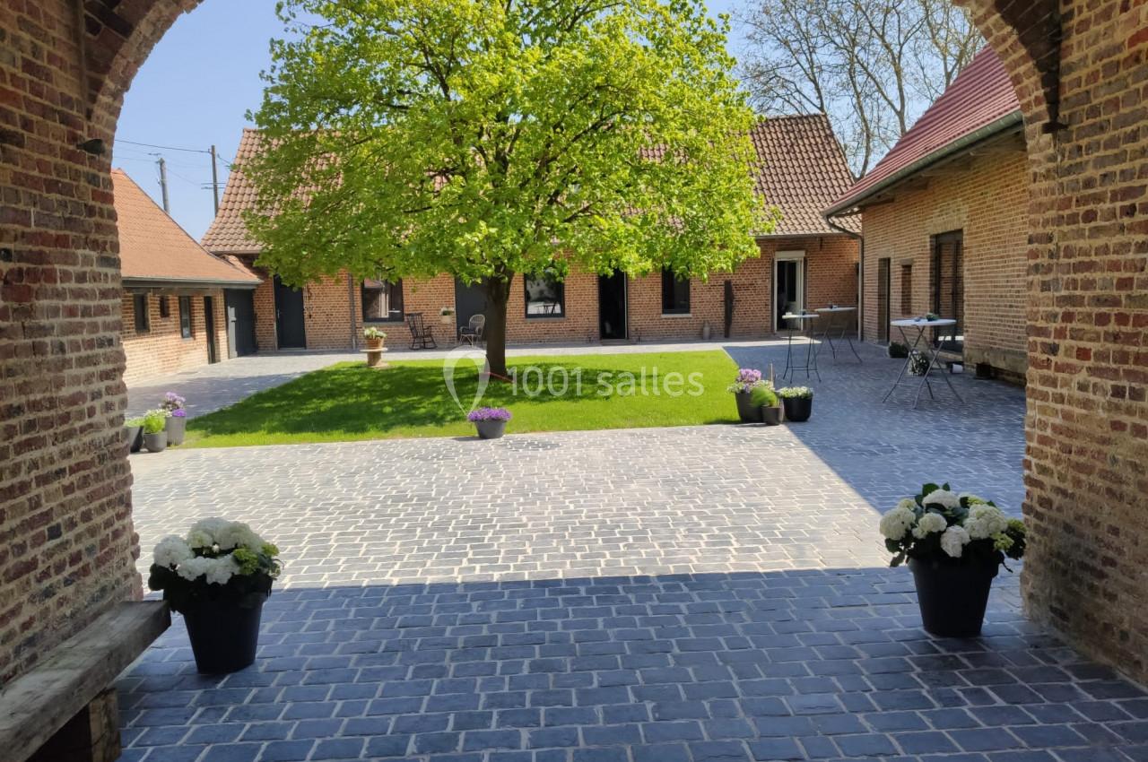 Location salle Haverskerque (Nord) - Ferme d'Hercule #35 Cour pavée avec un arbre central, entourée de bâtiments en briques, pots de fleurs et mobilier de jardin.
