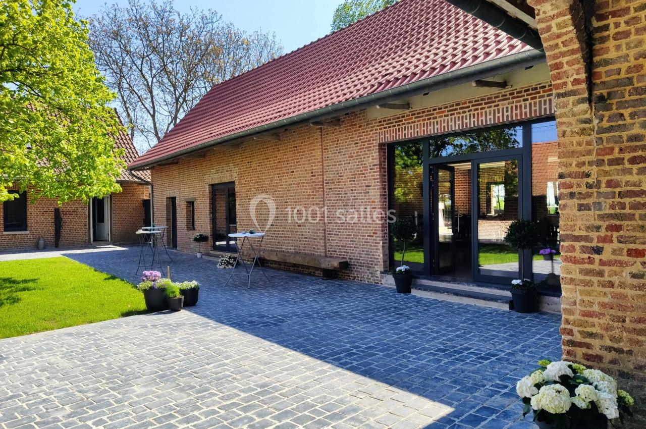 Location salle Haverskerque (Nord) - Ferme d'Hercule #36 Cour pavée avec bâtiment en briques rouges, grandes fenêtres et plantes en pots, entourée de verdure.