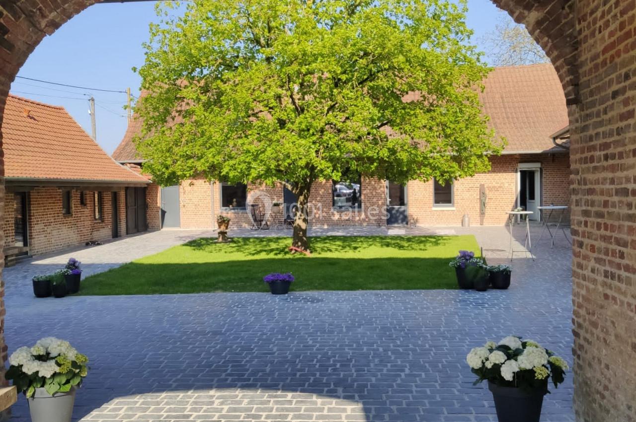 Location salle Haverskerque (Nord) - Ferme d'Hercule #5 Vue d'une cour pavée avec un arbre central, entourée de bâtiments en briques et décorée de pots de fleurs.