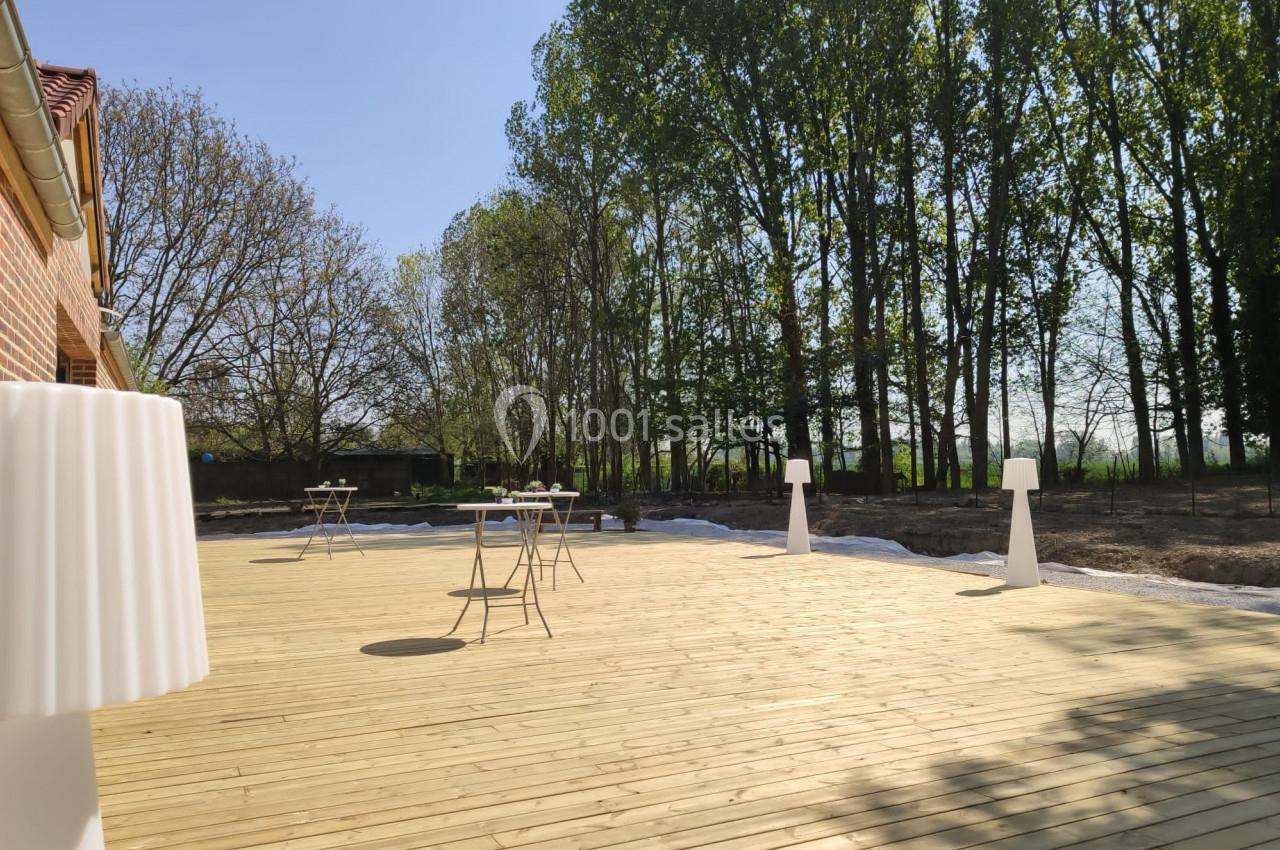 Location salle Haverskerque (Nord) - Ferme d'Hercule #26 Terrasse en bois avec quelques tables et lampes, bordée d'arbres sous un ciel dégagé.