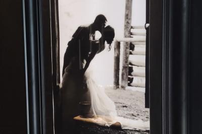 Miniature Location salle Haverskerque (Nord) - Ferme d'Hercule #37 Photographie en noir et blanc d'un couple s'embrassant, la femme en robe de mariée, devant une structure en bois.
