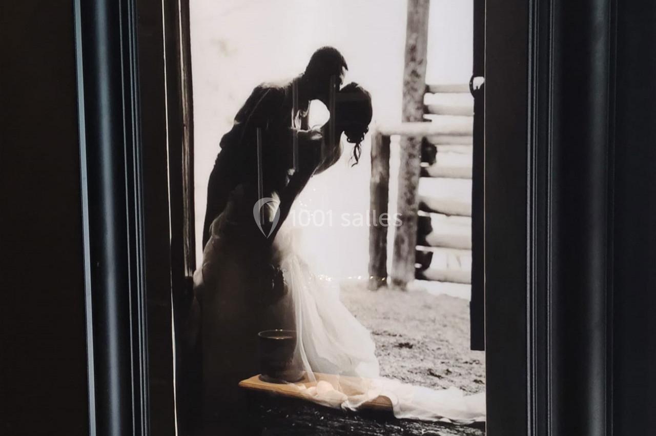 Location salle Haverskerque (Nord) - Ferme d'Hercule #37 Photographie en noir et blanc d'un couple s'embrassant, la femme en robe de mariée, devant une structure en bois.