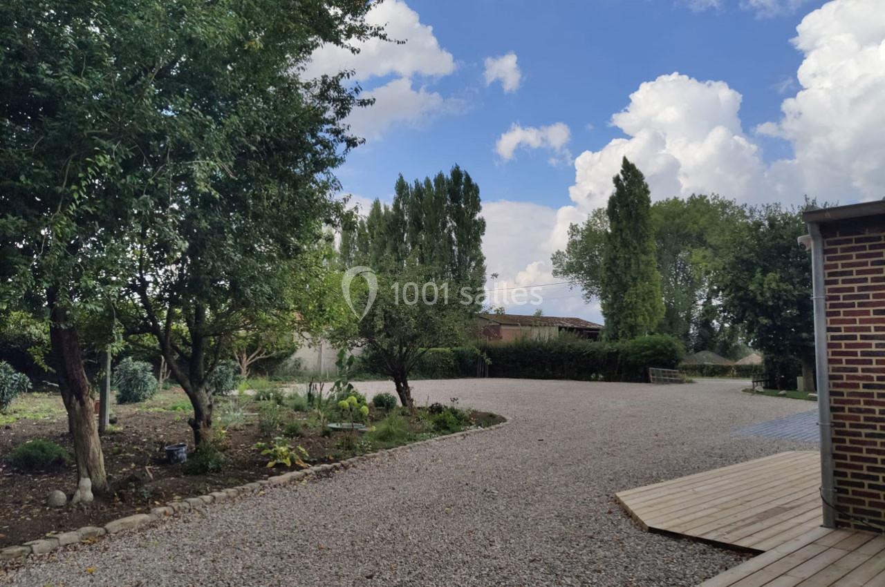 Location salle Haverskerque (Nord) - Ferme d'Hercule #24 Cour extérieure avec allée en gravier, bordée d'arbres et d'arbustes, et terrasse en bois à droite sous un ciel…