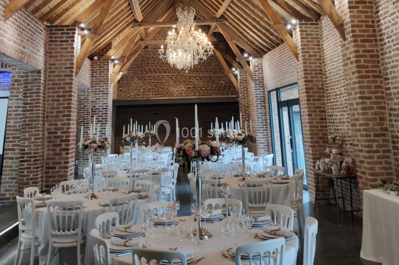 Location salle Haverskerque (Nord) - Ferme d'Hercule #2 Salle de réception avec murs en briques, poutres apparentes, tables rondes dressées et lustre central.