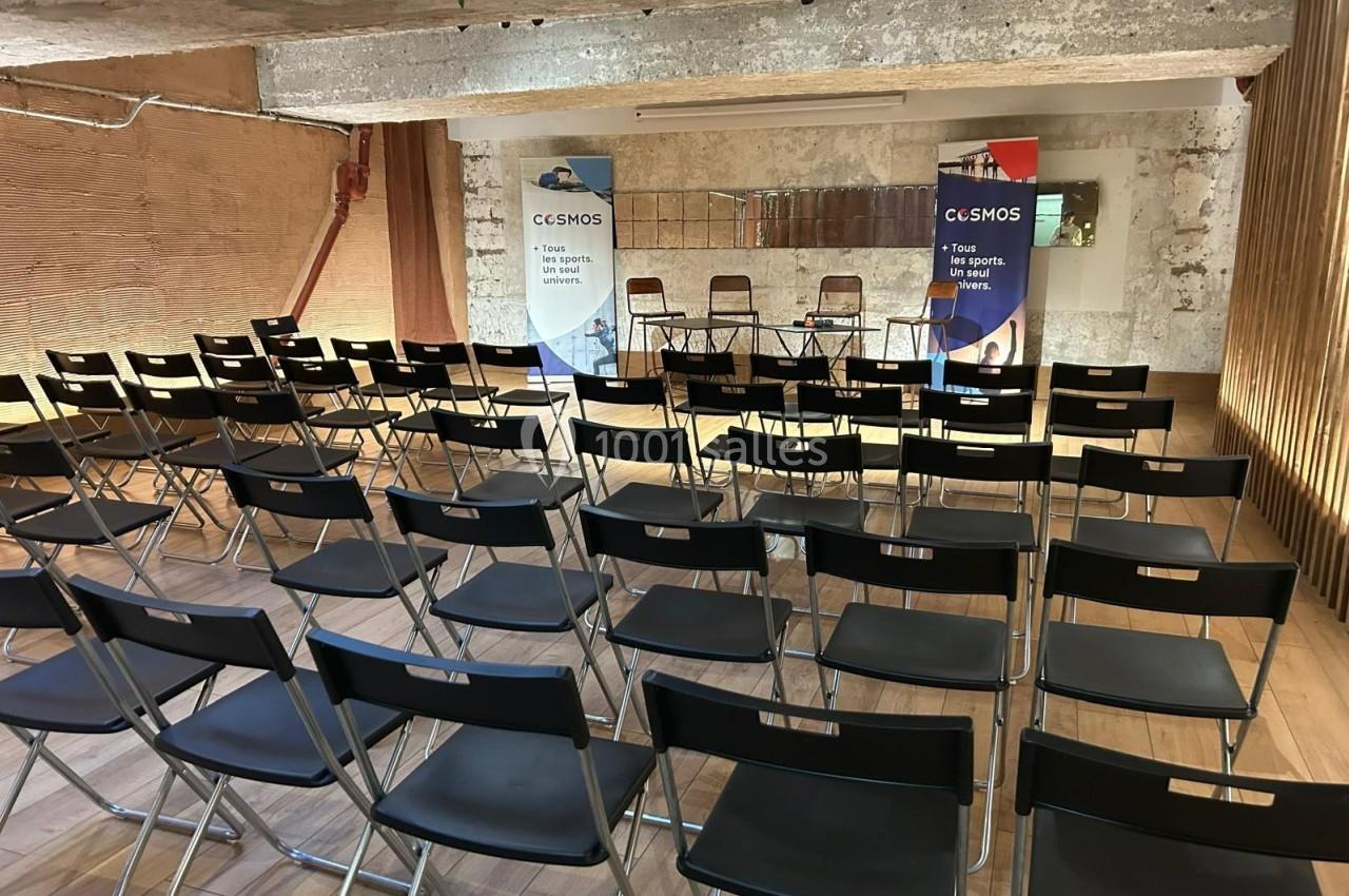 Salle de conférence vide avec des rangées de chaises noires pliantes et deux kakémonos colorés en arrière-plan.