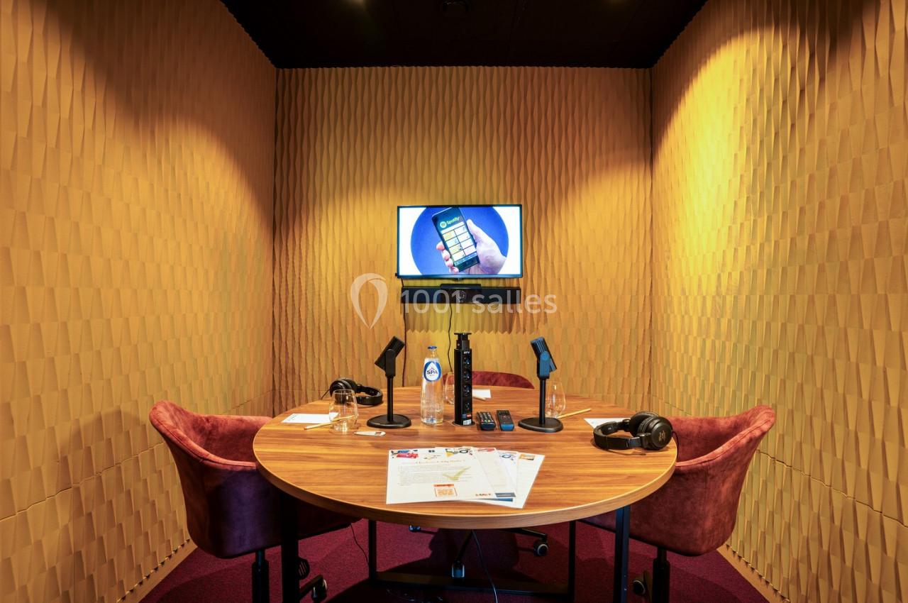 Salle de réunion isolée avec table ronde, chaises, microphones, casque audio, écran mural et documents posés.