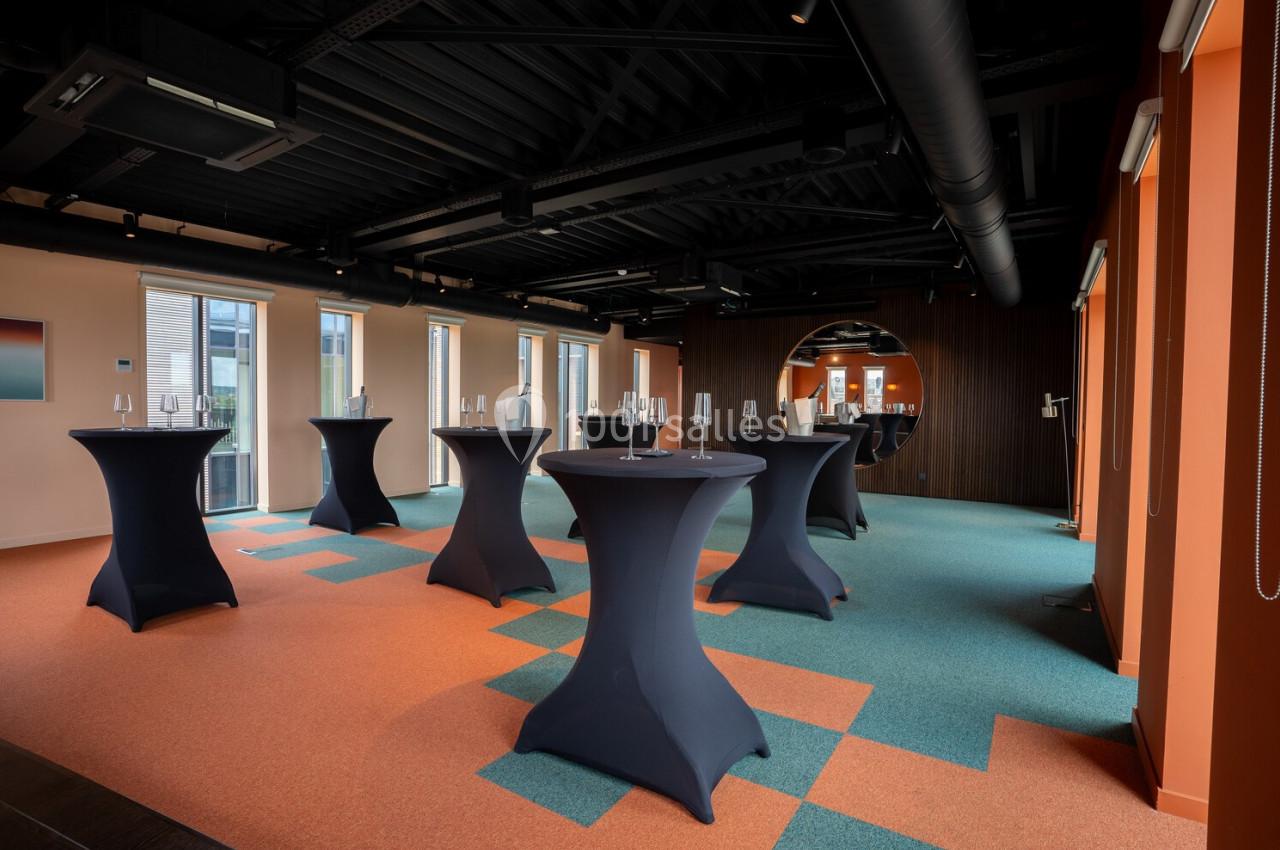 Salle moderne avec tables hautes noires, sol coloré orange et bleu, grandes fenêtres et éclairage naturel.