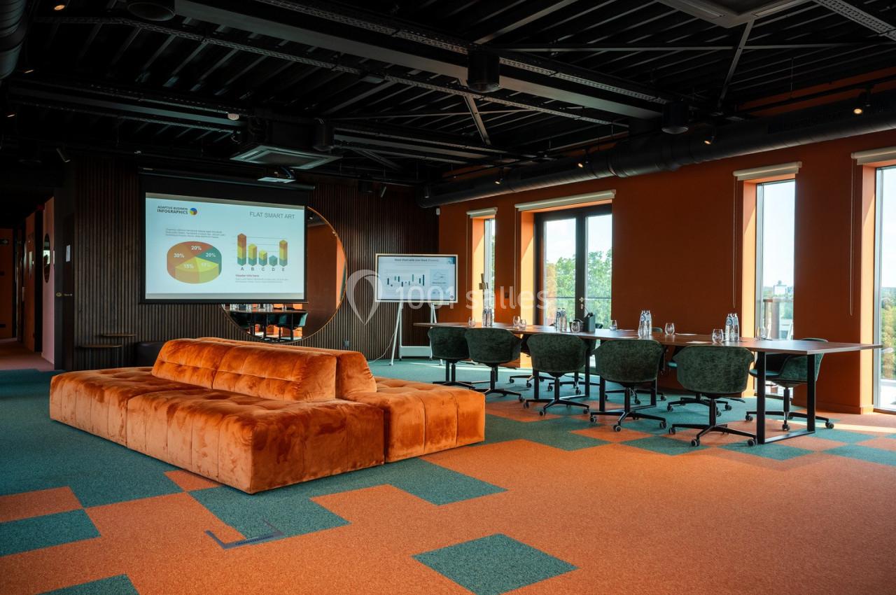 Salle de réunion moderne avec canapé orange, écran de présentation, tableau blanc et grandes fenêtres.