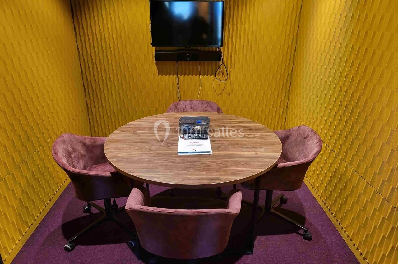Salle de réunion compacte avec murs jaunes texturés, table ronde en bois, quatre chaises roses et un écran mural.