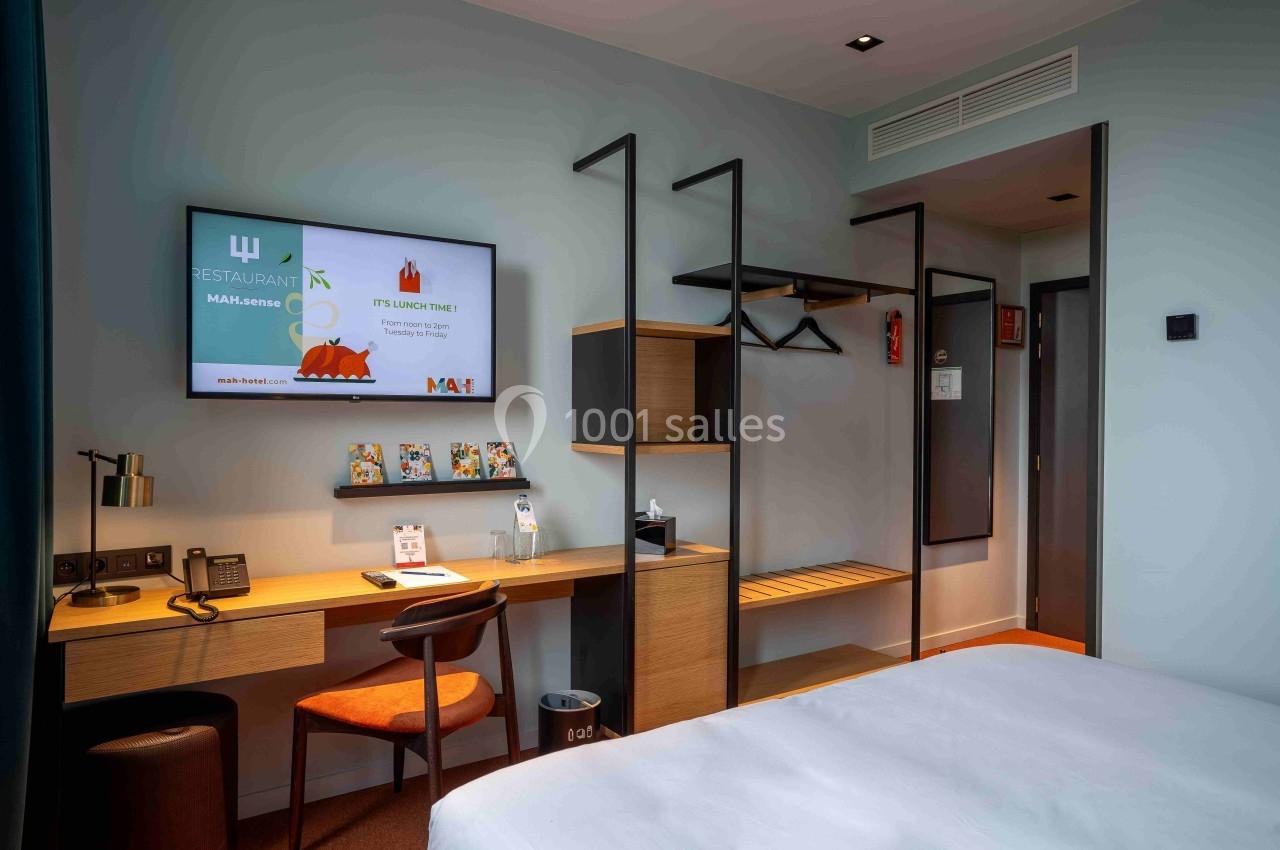 Chambre d'hôtel moderne avec bureau, téléviseur mural, étagères ouvertes et lit partiellement visible au premier plan.