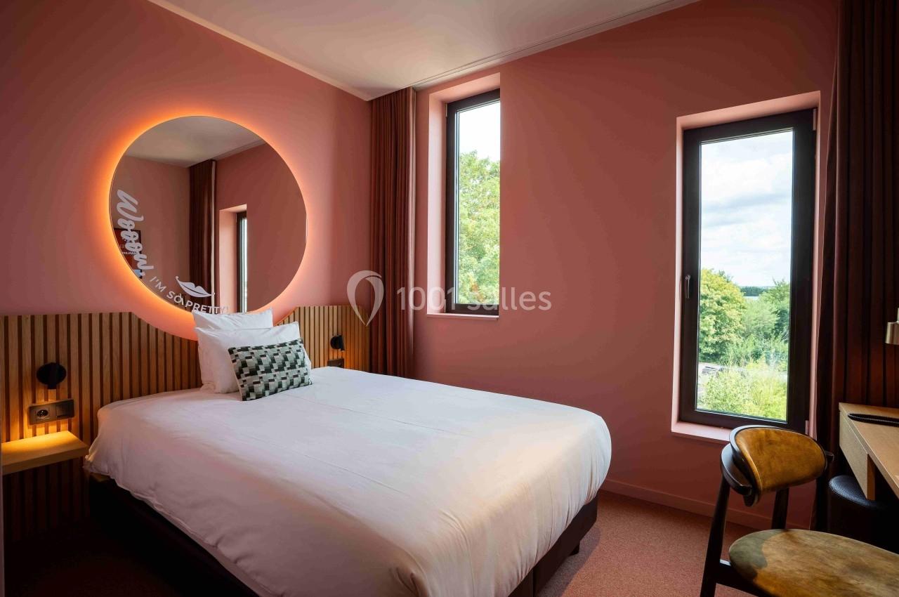 Chambre d'hôtel avec lit double, mur rose, miroir rond éclairé, bureau, chaise et fenêtres donnant sur un paysage verdoyant.