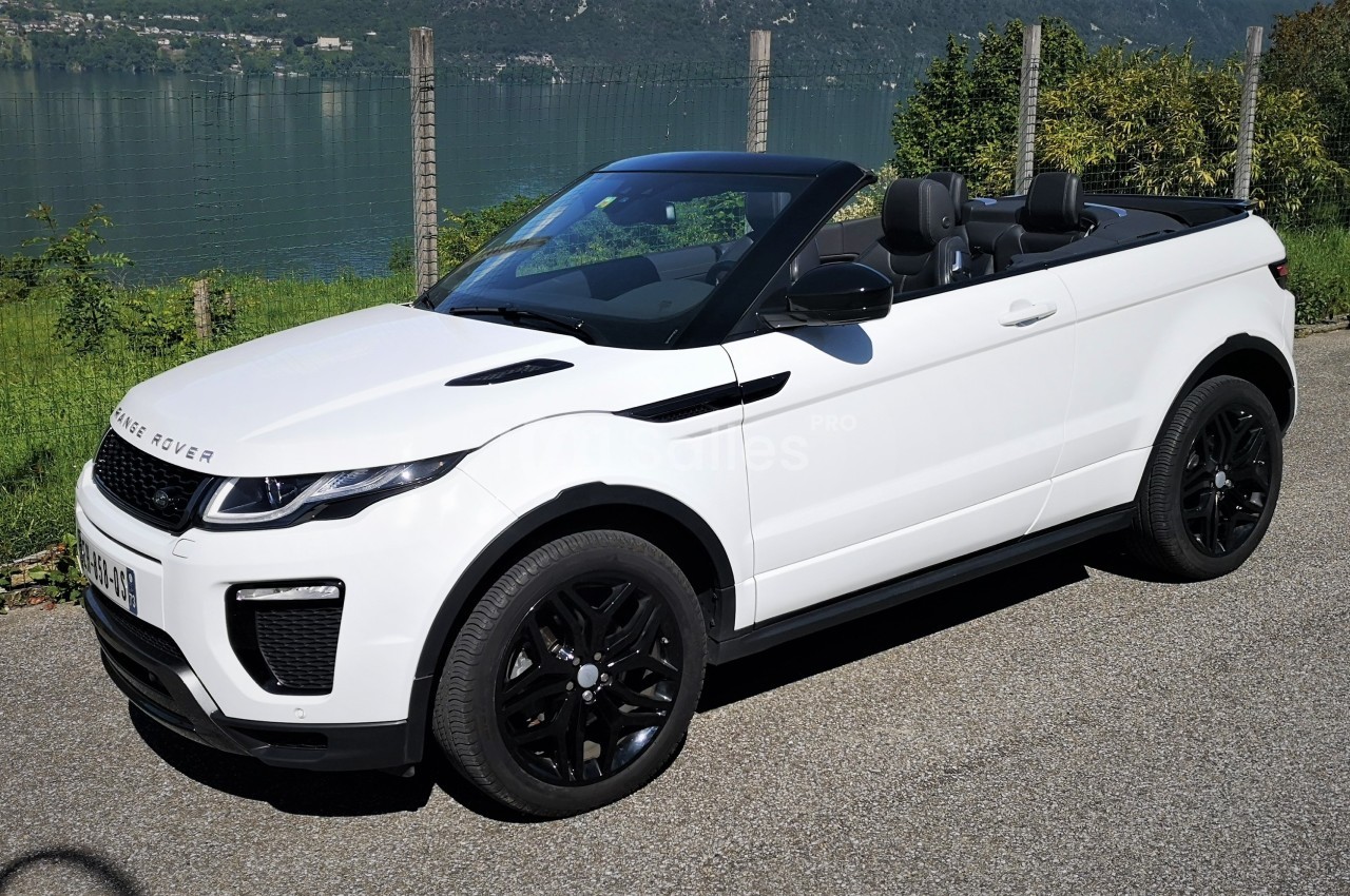 Voiture décapotable blanche stationnée sur une route, avec un lac et des collines en arrière-plan.