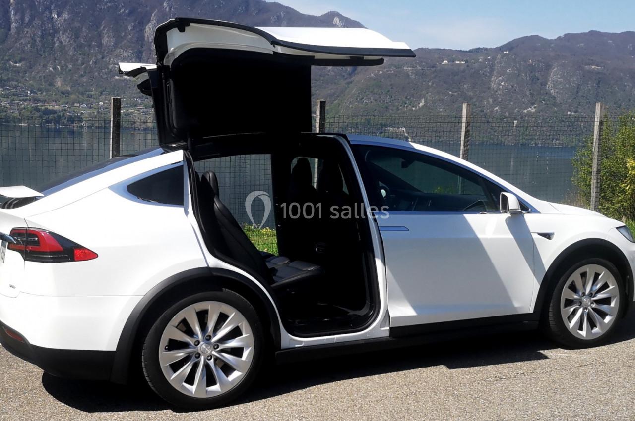 Voiture blanche avec portes arrière en ailes de mouette ouvertes, stationnée près d'un lac et de montagnes.