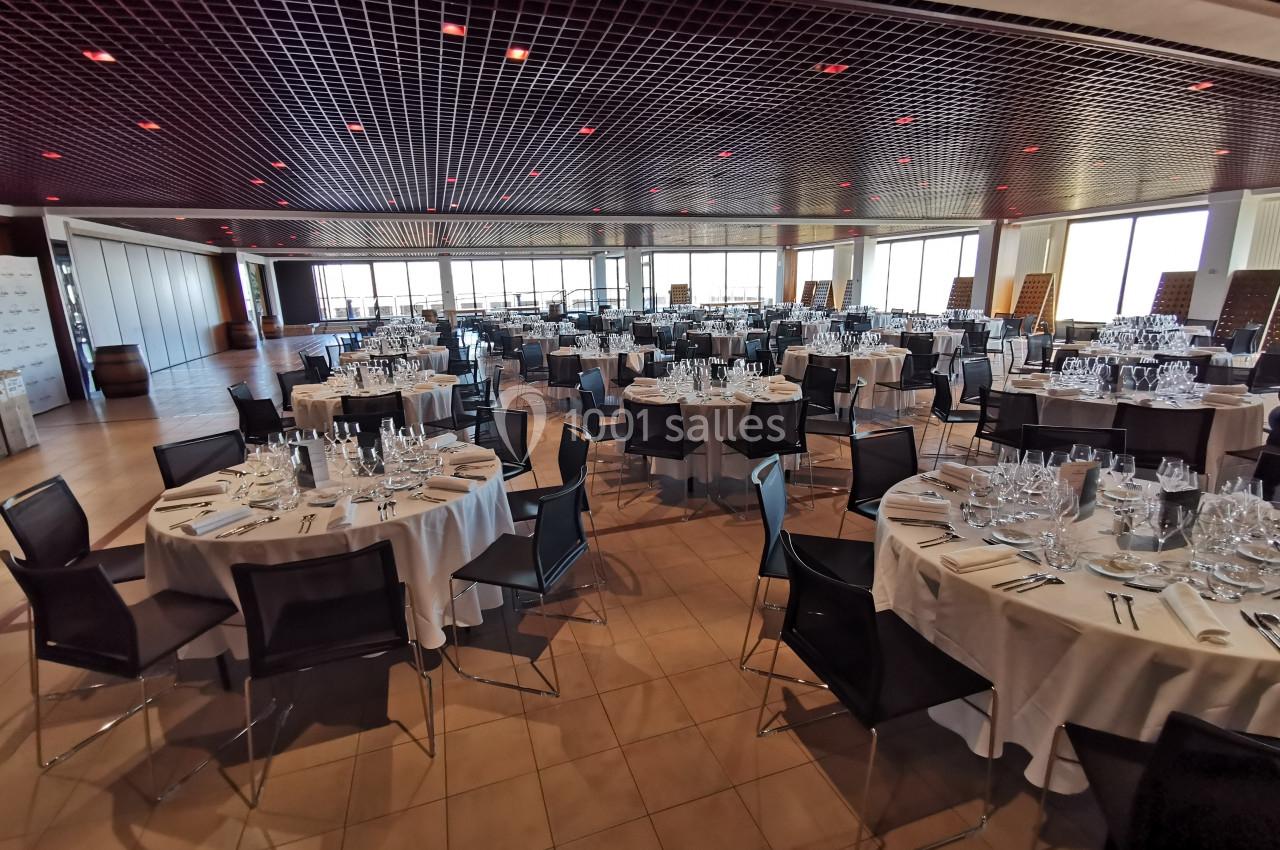 Salle de réception avec des tables rondes dressées, nappes blanches, chaises noires et lumière naturelle.