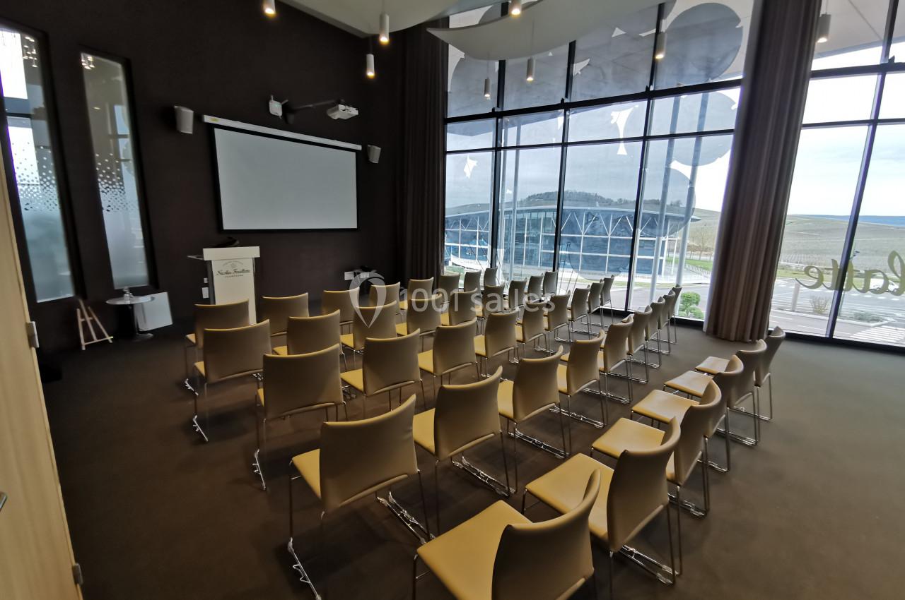 Salle de conférence moderne avec chaises alignées, écran de projection et grandes baies vitrées donnant sur l'extérieur.