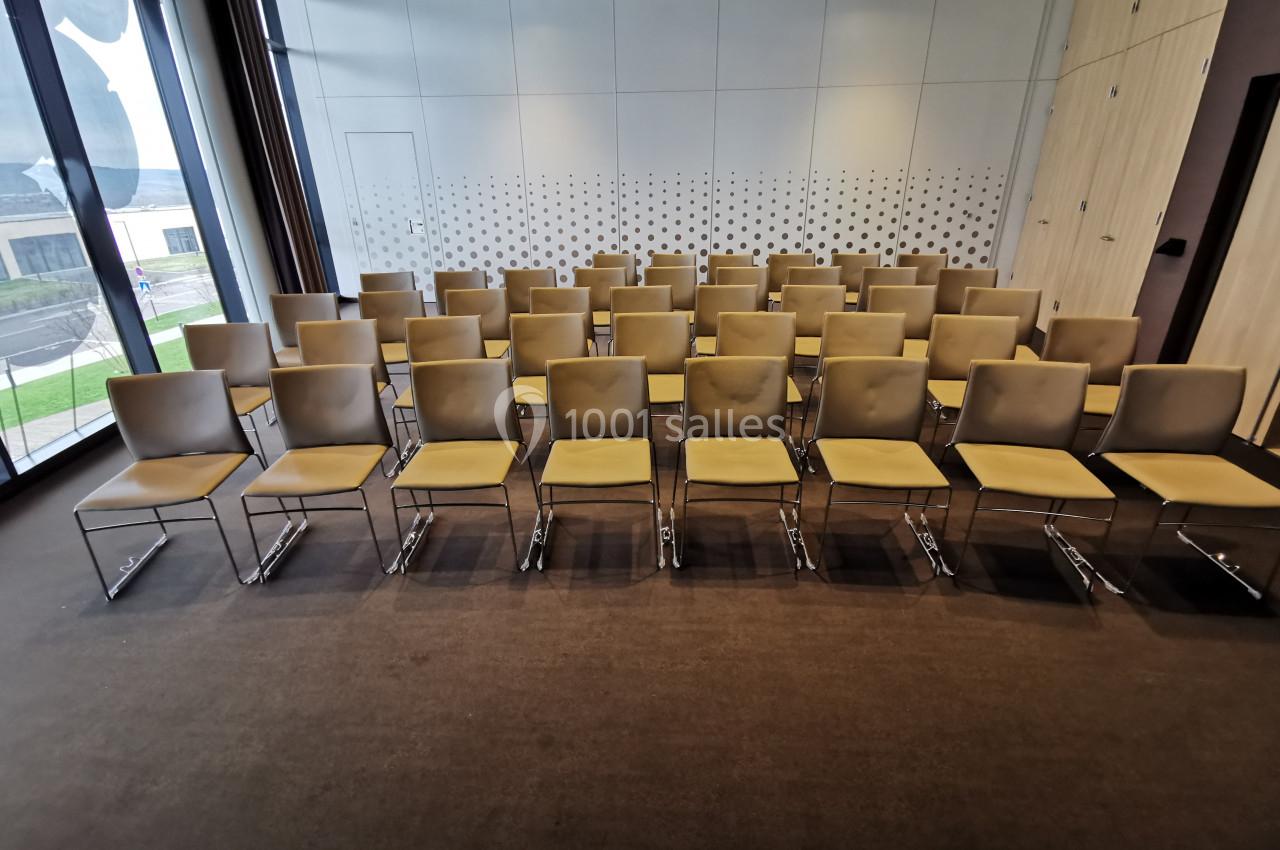Salle de réunion avec des rangées de chaises beige alignées sur un sol marron, devant un mur vitré et perforé.