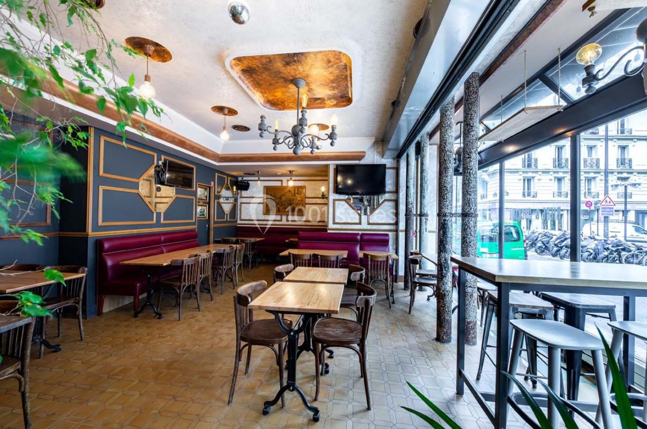 Salle de restaurant lumineuse avec tables en bois, banquettes rouges, miroirs muraux et grandes fenêtres donnant sur la rue.