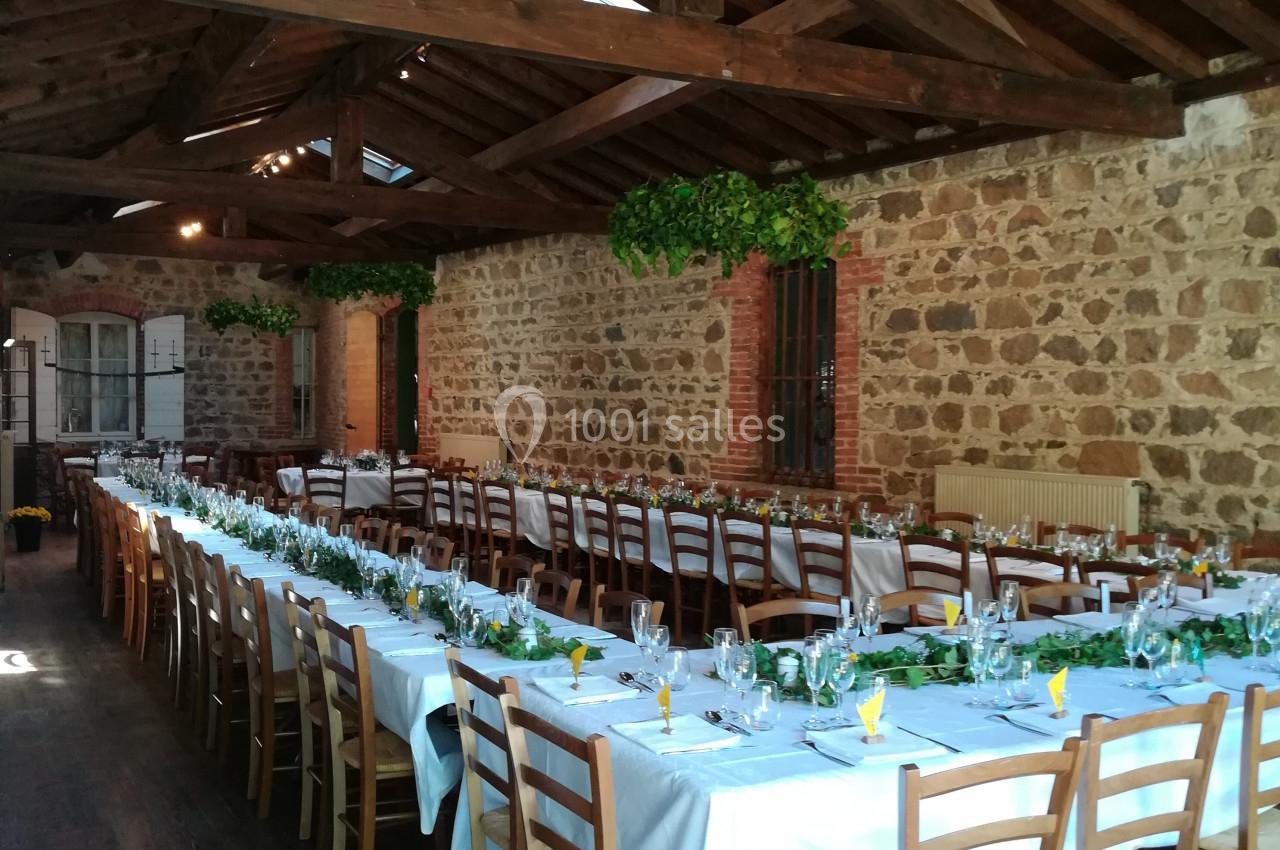 Salle en pierre avec plafond en bois, tables longues dressées avec nappes blanches, chaises en bois et décoration végétale.